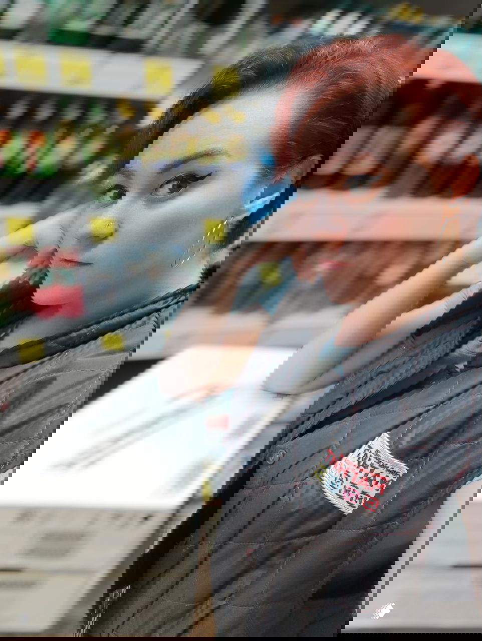 Портретная фотография сотрудницы в униформе магазина «Магнит» на фоне полок с напитками, лицо и жилет с бейджем видны крупным планом.