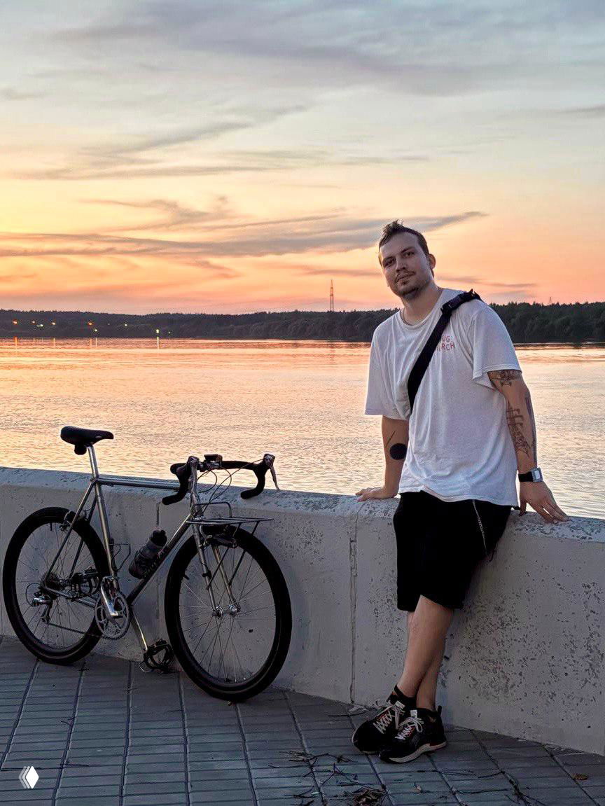 Человек стоит у набережной рядом с шоссейным велосипедом на фоне заката над рекой — городской портрет с велосипедом и вечерним небом.