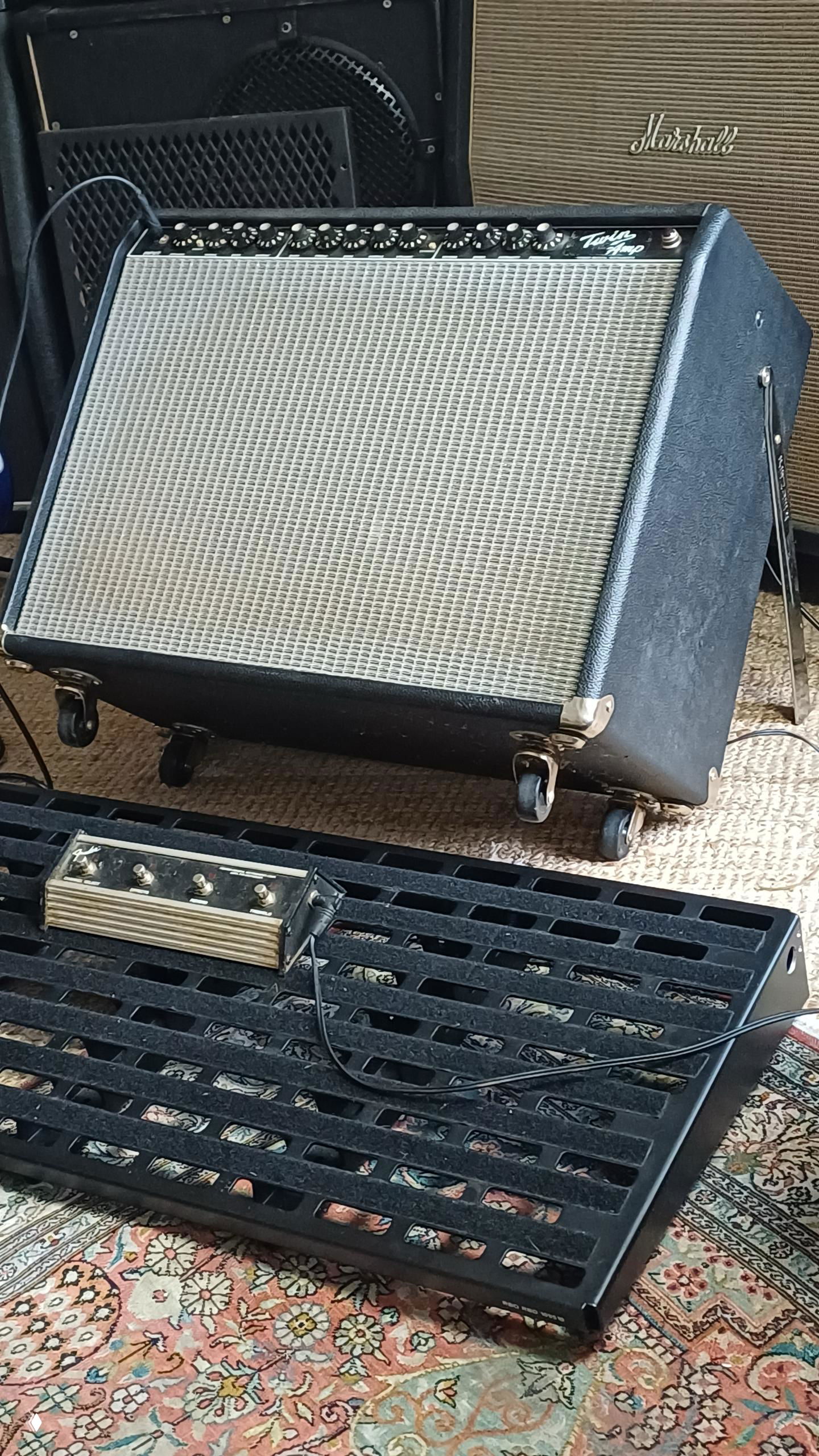 Ламповый усилитель на колесиках и педалборд на ковре: видна решётка динамика, панель с регуляторами и подключённые кабели в мастерской.