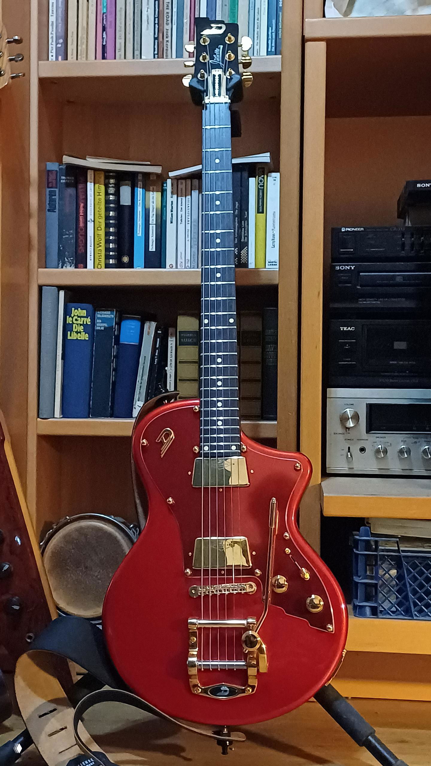 Красная электрогитара с золотой фурнитурой на фоне книжного шкафа и стеллажей, фото найденного инструмента в коллекции.