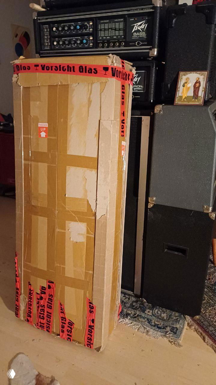 Высокая узкая коробка, усиленная скотчем и лентой 'Vorsicht Glas', стоит возле усилителей и музыкальной аппаратуры — упаковка музыкальных инструментов.