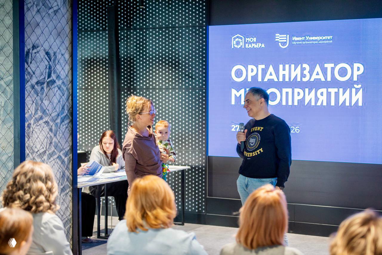 Общение с участницей курса, мужчина с микрофоном беседует с женщиной, в руках у неё ребёнок — тёплая сценка после выступления.