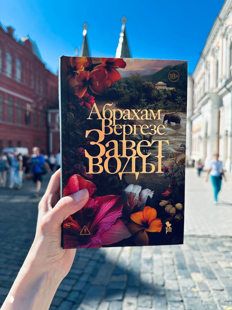 Крупный план книги в руке: новое издание Абрахама Вергезе «Завет воды» на фоне размытых зданий и городской сцены после фестиваля.