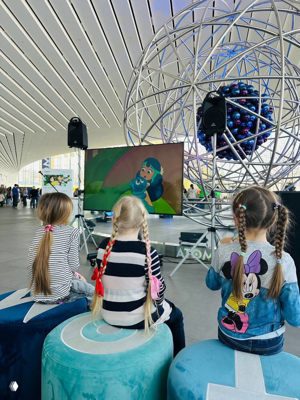 Дети смотрят экран с короткой анимацией в общей зоне фестиваля: три девочки с косами сидят на пуфах перед большим монитором и декоративной инсталляцией.