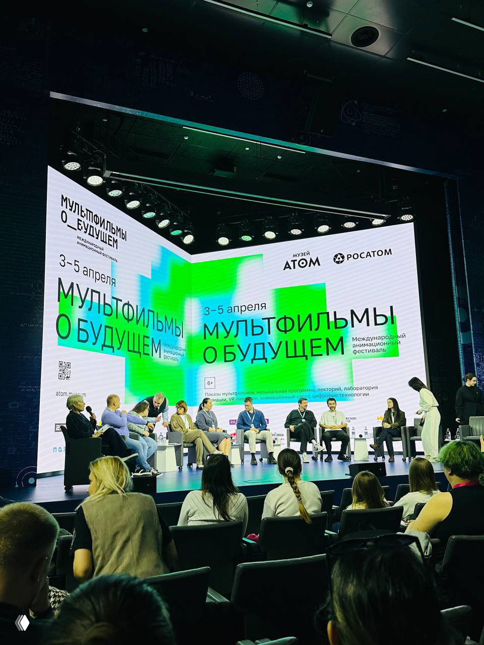 Панорамный кадр с конференц-панелью фестиваля: сцена с участниками, большой экран с постером «Мультфильмы о будущем» и аудитория слушателей в первом ряду.