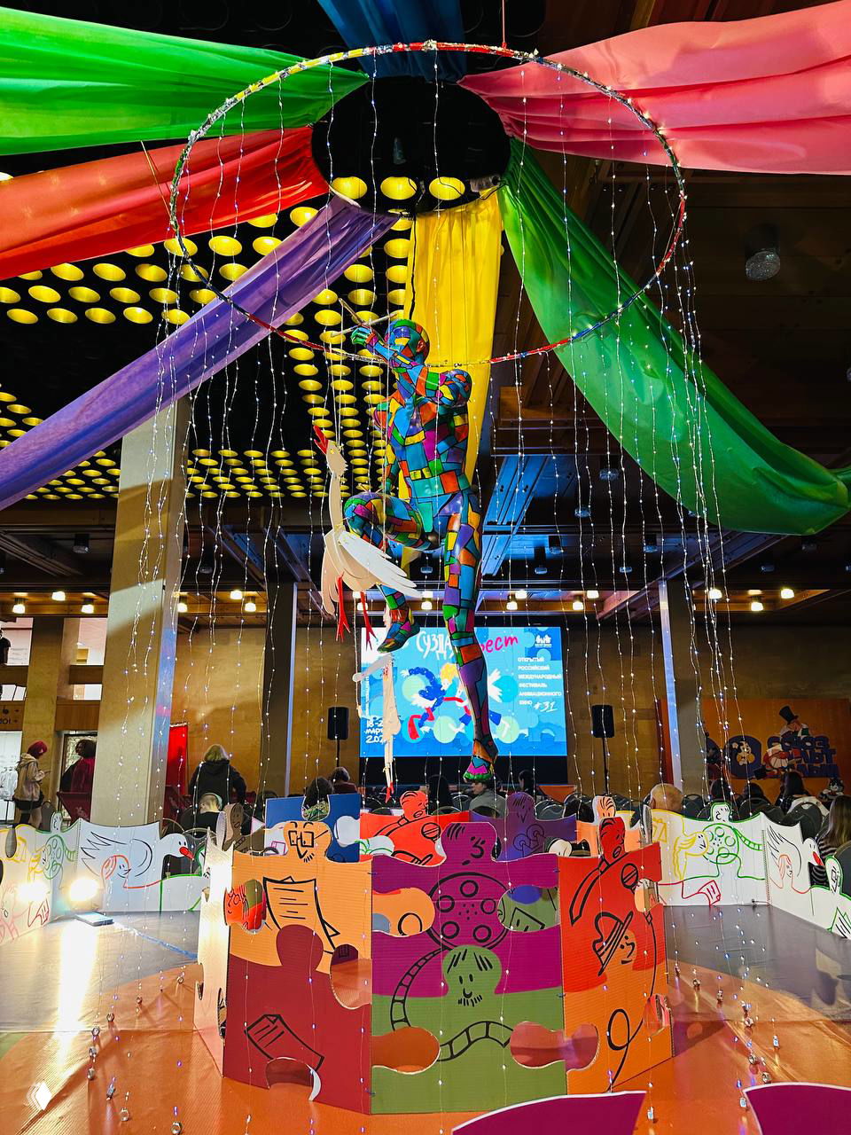 Яркая интерьерная сцена с цветной театральной инсталляцией и подвесной фигурой, декоративные ткани и огни на фоне зала фестиваля.