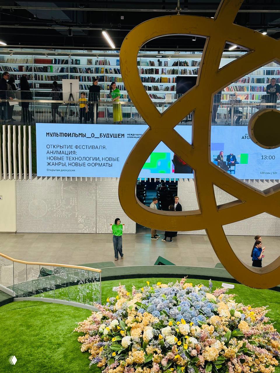 Интерьер площадки фестиваля: крупный декоративный символ на переднем плане, цифровой экран с информацией о программе и цветочная инсталляция внизу.