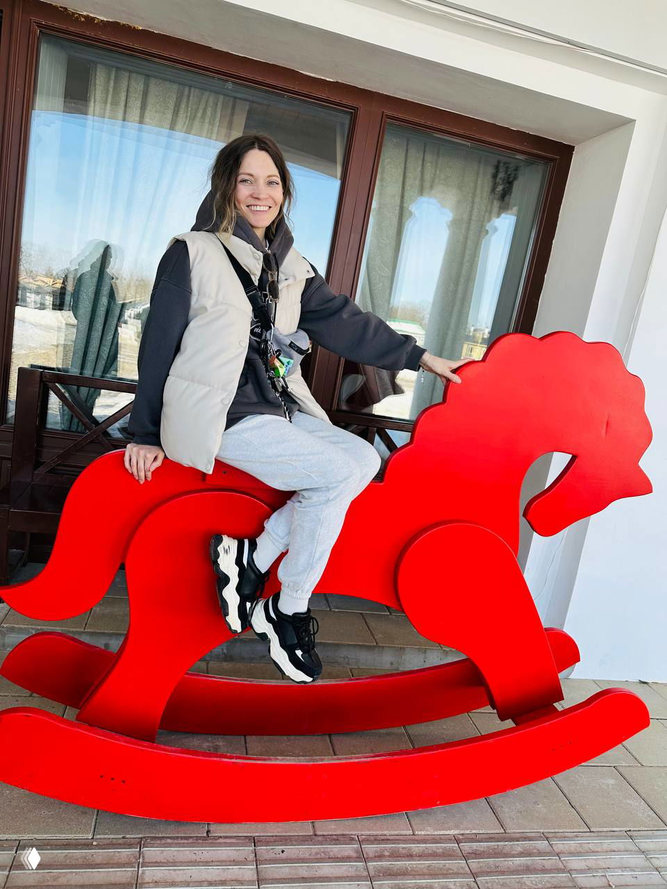 Женщина улыбается, сидя на большой красной детской качалке у входа; кадр передаёт непринуждённую атмосферу поездки и отдыха.