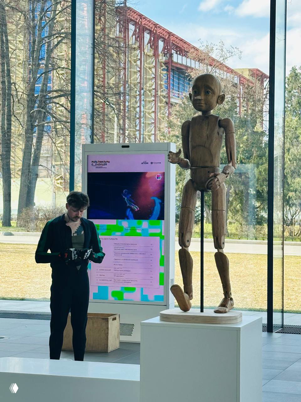 Экспонат и перформанс на фоне панорамного окна: деревянная кукла-манекен на постаменте и человек в перчатках рядом, видна внешняя архитектура за стеклом.