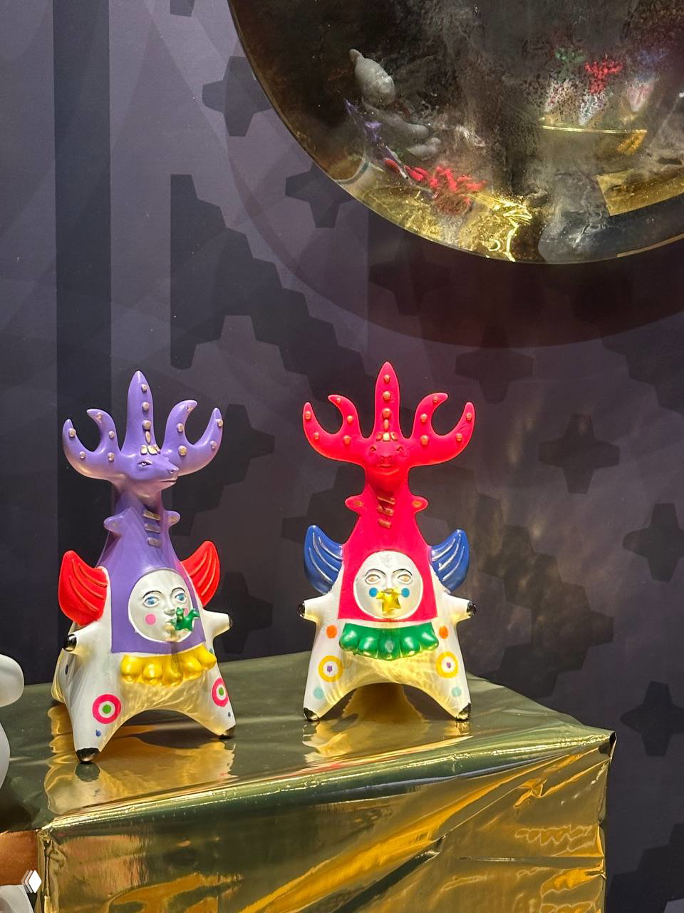 Яркие авторские игрушки с сюрреалистичными формами: парные фигурки с декоративными рожками и расписными лицами на блестящей подставке.