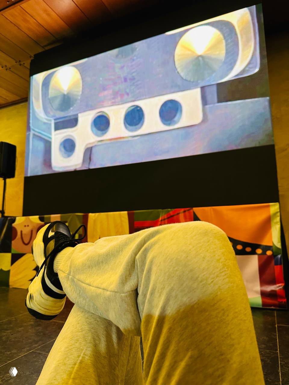 Ноги в кроссовках на переднем плане и экран с проекцией на фоне — кадр из зала, где показывают фильм или презентацию для семейной аудитории.