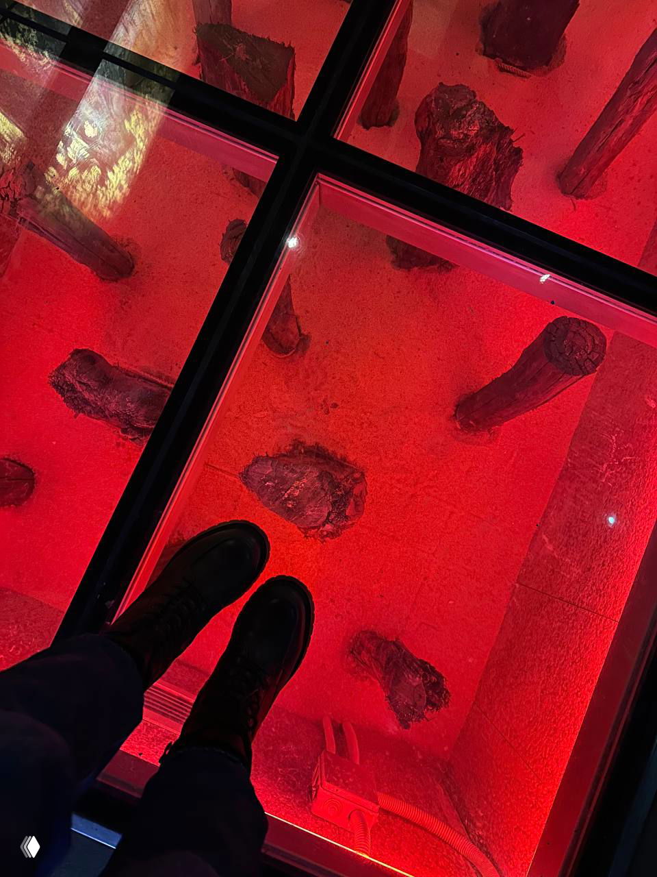 Стеклянный пол экспозиции с подсветкой красного цвета, под которым видны археологические или декоративные предметы — фотография с ногами автора на переднем плане.