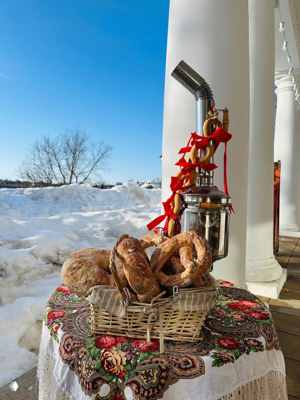 Корзина с бубликами на павильоне рядом с самоваром, украшенным ленточками, снег за колоннами и традиционная русская скатерть под подносом.