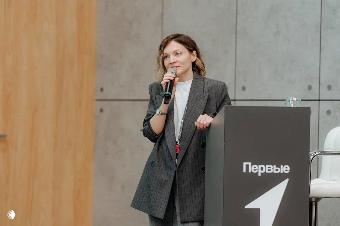 Спикер у подиума с микрофоном в руке на фоне брендинга мероприятия; образ в деловом пиджаке, формат доклада на сцене.