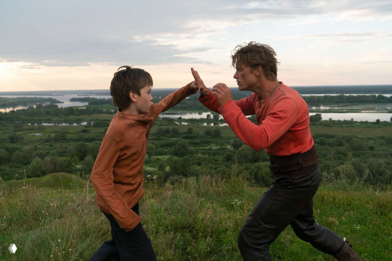 Сцена тренировки на природе: подросток и взрослый мужчина отрабатывают приемы на холме с рекой и зеленью в глубине кадра