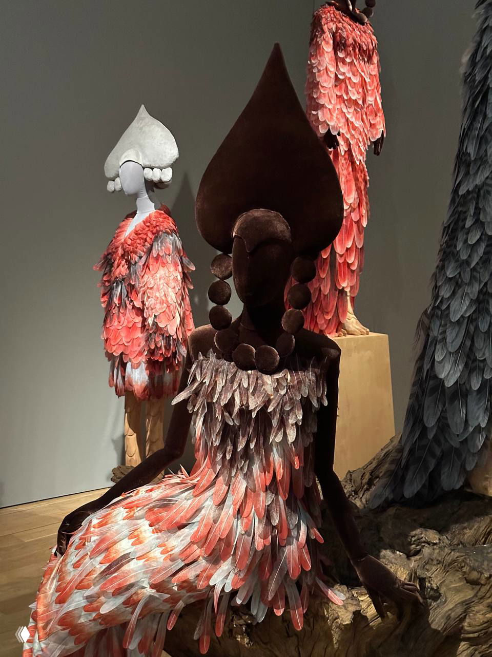 Манекен в платье с перьевой или чешуйчатой фактурой розово-красных тонов на фоне других фигур, создающих образ сказочного леса — цвет и фактура.