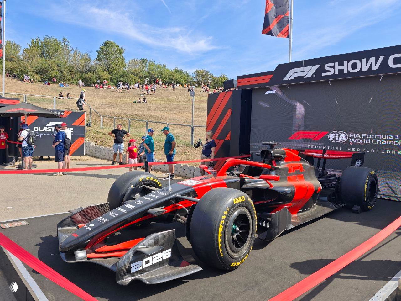 Красный демонстрационный болид Formula 1 на подиуме Show Car с ограждениями и посетителями вокруг; крупный план шасси и передней части машины в солнечный день.