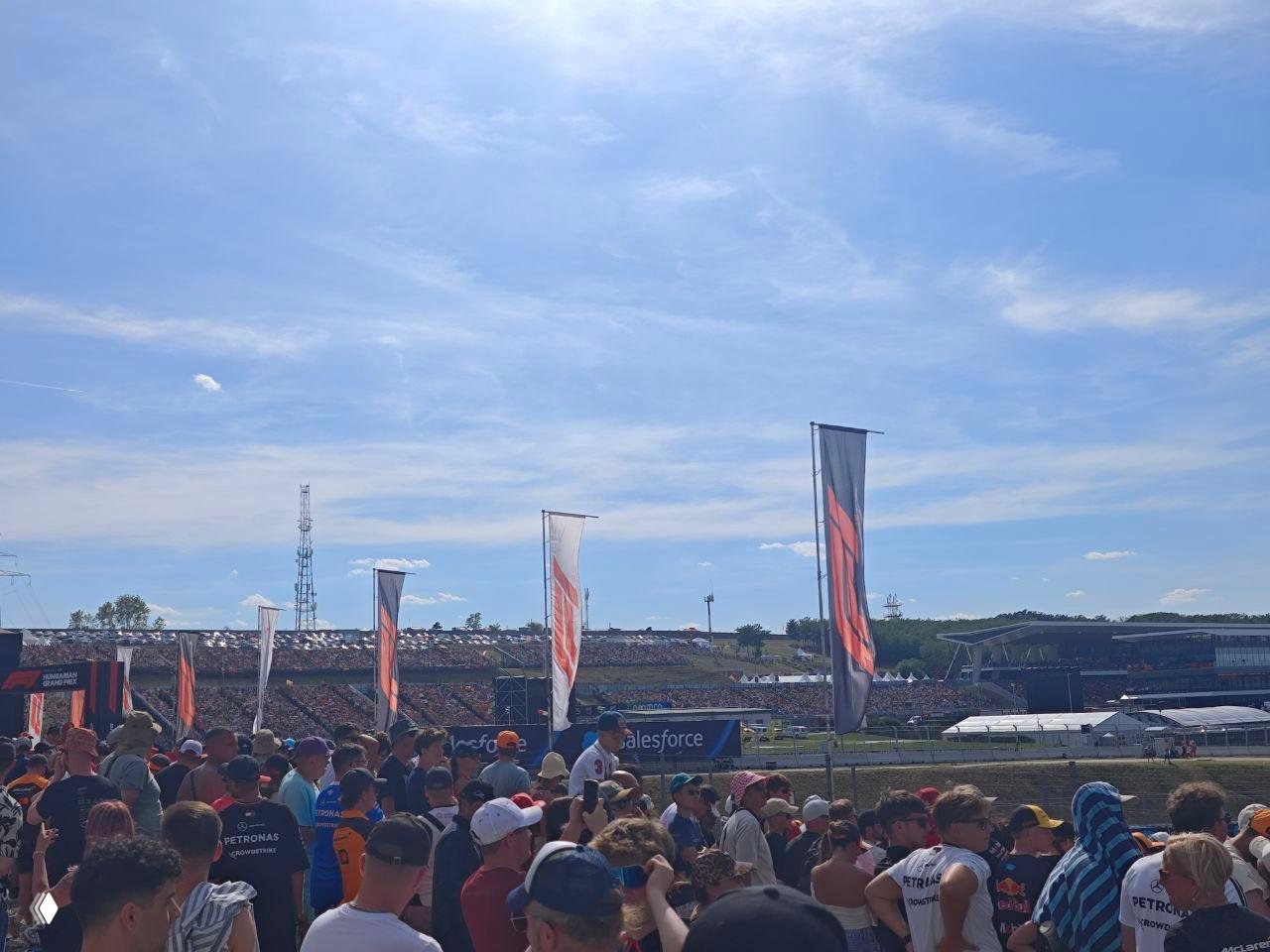 Толпа зрителей у барьера трассы на фоне трибун и холмов Hungaroring в ясный летний день; люди смотрят заезда и создают фестивальную атмосферу уик‑энда.