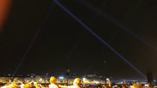 Вид с площадки на ночной Будапешт: лазерные лучи, городские огни и толпа людей, наблюдающих за праздничной программой и салютом.