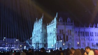 Подсвеченный синим фасад здания парламента в Будапеште вечером, на переднем плане — толпы людей и праздничная атмосфера вокруг.