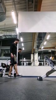 Тренажёрный зал: мужчина в спортивной форме идёт рядом со штангой и скамьёй, отражение в зеркале, кадр перед началом упражнения и подготовки в зале.