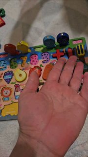 Рука ребёнка рядом с деревянным сортером с цифрами и цветными фигурками; кадр домашнего видео, демонстрирующий развивающую игру и похвалу.