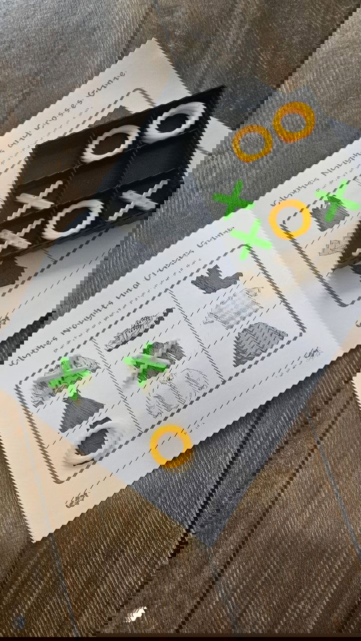 Распечатанное поле и пластиковые фишки крестик и нолик зелёного и жёлтого цветов, расположенные в коробочке на деревянном столе рядом с листом игры.