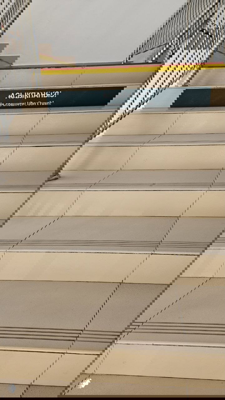 Лестница в поликлинике Алматы с табличкой «Асықпаңыз! Не спешите! Don't rush!», видны перила и ступени.