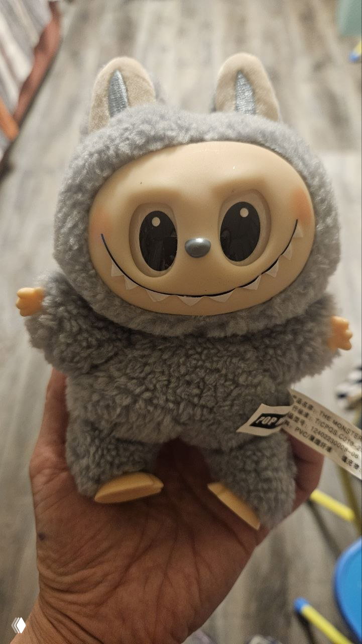 Игрушка Labubu серого цвета в руке взрослого, мягкий ворс и выразительное лицо персонажа, детская коллекционная игрушка.