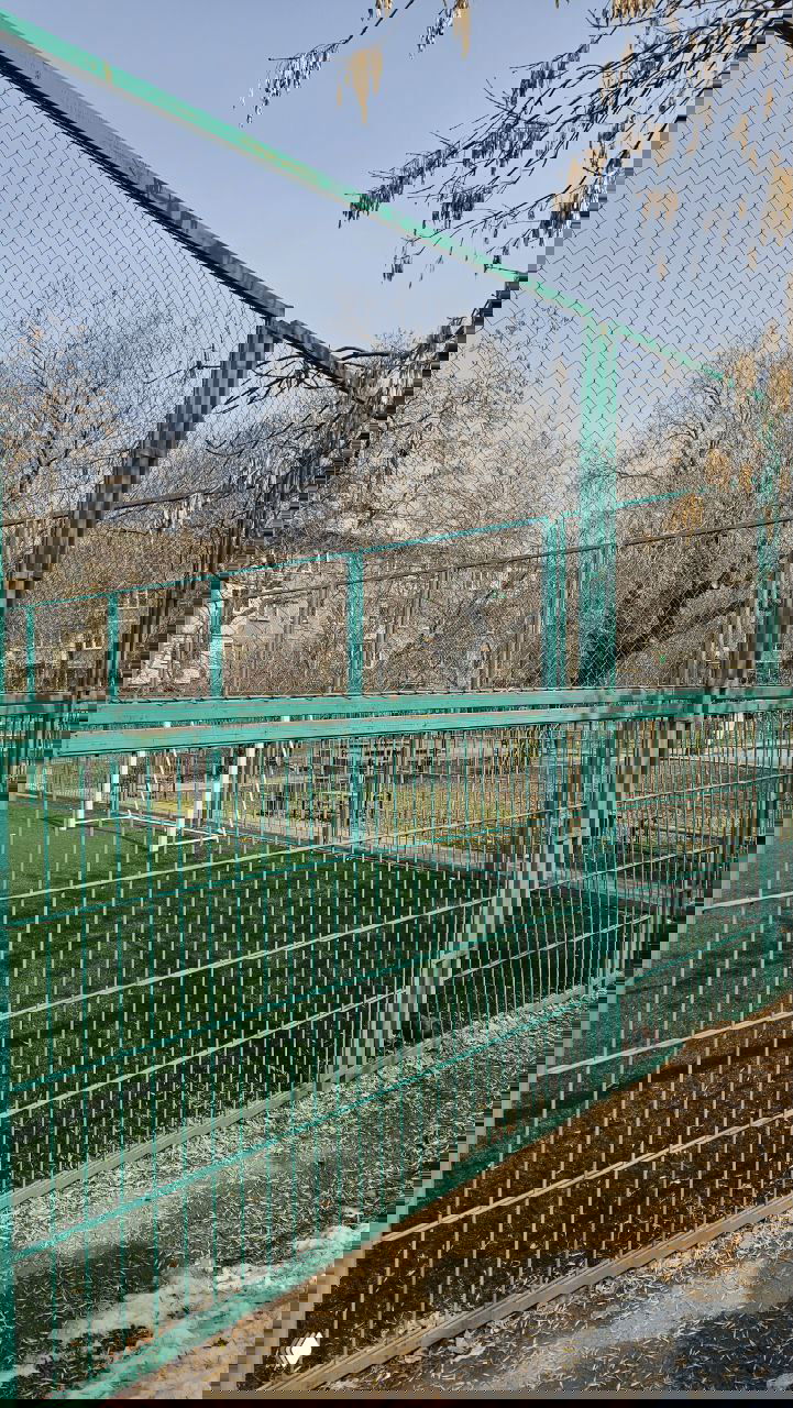 Крупный план зелёного металлического ограждения дворового футбольного поля, видна часть искусственной травы и деревья вокруг.