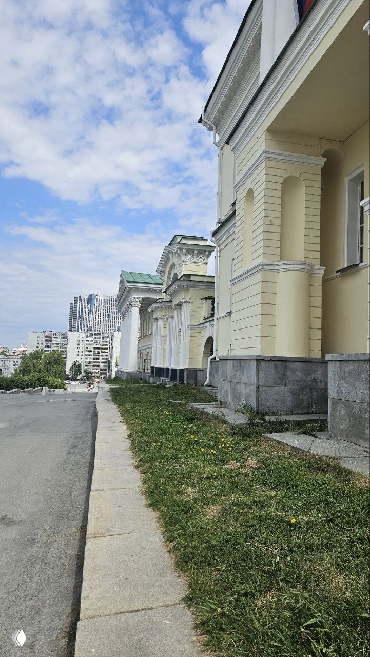 Ряд светло‑жёлтых классических фасадов здания у тротуара, передний план газон и городской силуэт высоток на фоне голубого неба.
