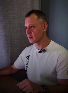 Мужчина в белой футболке с прищепленным петличным микрофоном, сидит за столом в кабинете и говорит, глядя в сторону экрана в нейтральном интерьере.