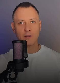 Крупный план мужчины в студии: говорит в микрофон на нейтральном фоне, видны лицо, микрофон и верхняя часть торса.