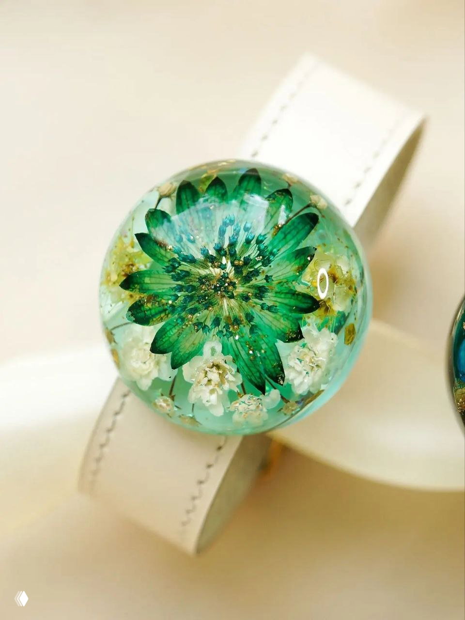 Крупный план круглого кабошона из прозрачной смолы с зелёным цветком и маленькими белыми бутонами на светлом ремешке.