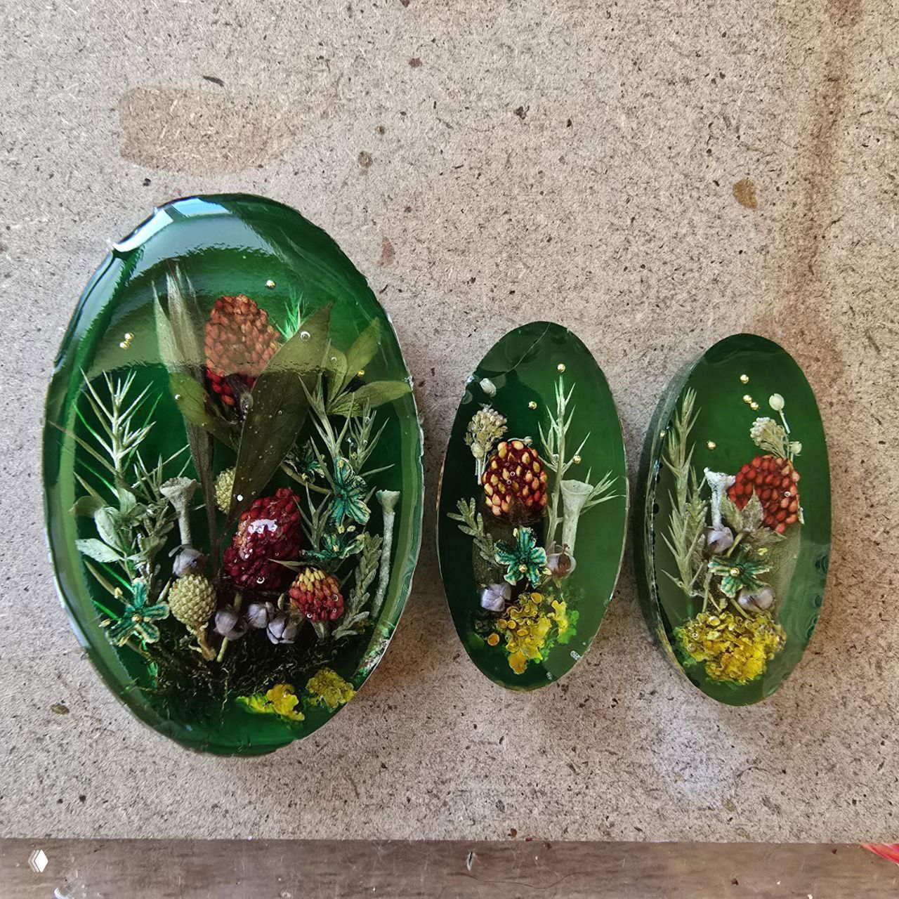 Три овальных кабошона ярко‑зеленого цвета с композициями из шишек и сухих растений в заливке эпоксидной смолы.