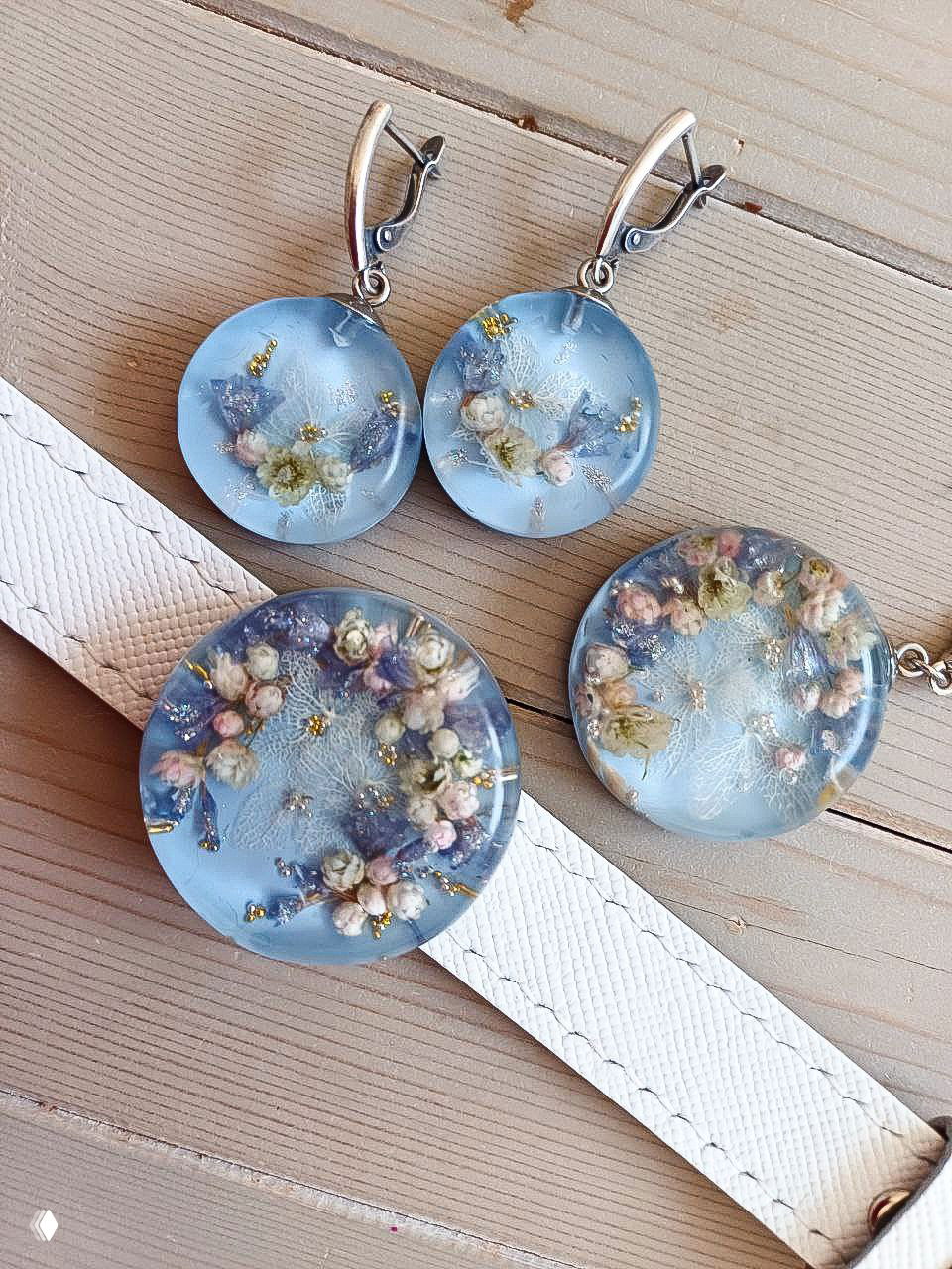 Набор круглых серёг и медальона в пастельно‑голубых тонах с мелкими белыми цветами и золотыми акцентами в смоле.