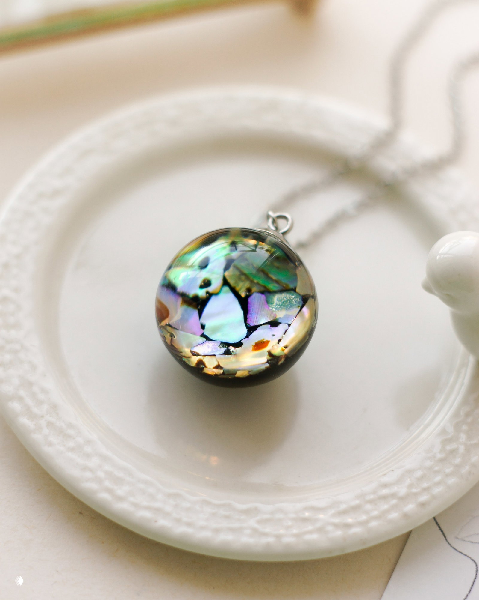 Круглый кулон с инкрустацией перламутром на белой тарелке, демонстрирует вариант исполнения вставок и текстуры поверхности готового изделия.