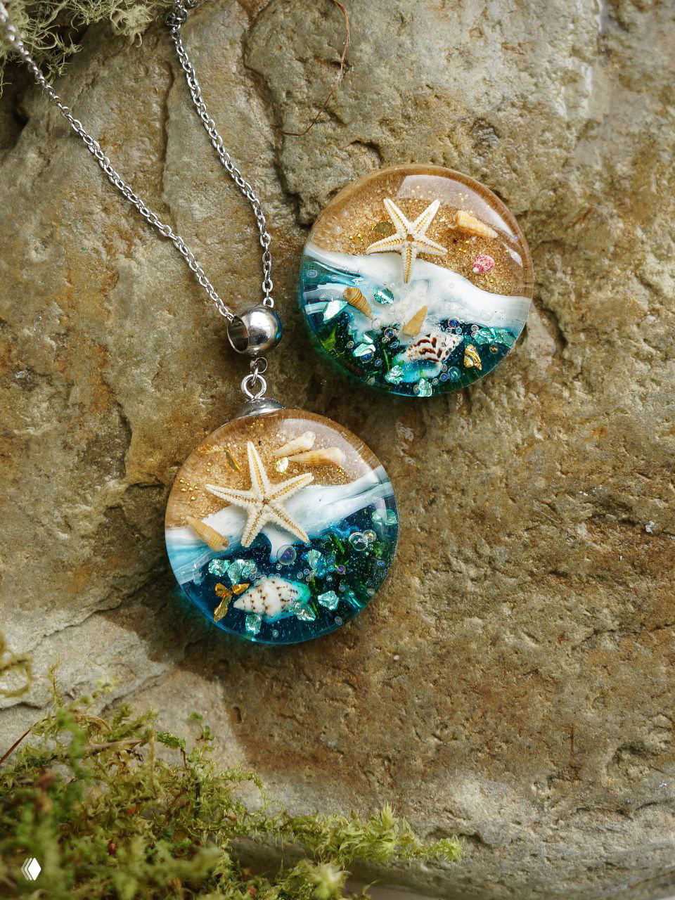 Две круглые подвески в морском стиле: звёздочки, ракушки и блёстки внутри прозрачной смолы на каменном фоне рядом с цепочкой.