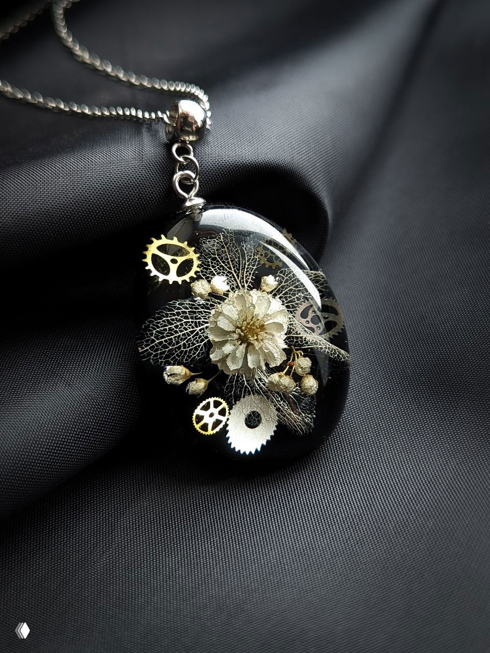 Овальная черная подвеска с белым цветком и металлическими шестерёнками внутри прозрачной смолы, контрастная предметная постановка на чёрном фоне.