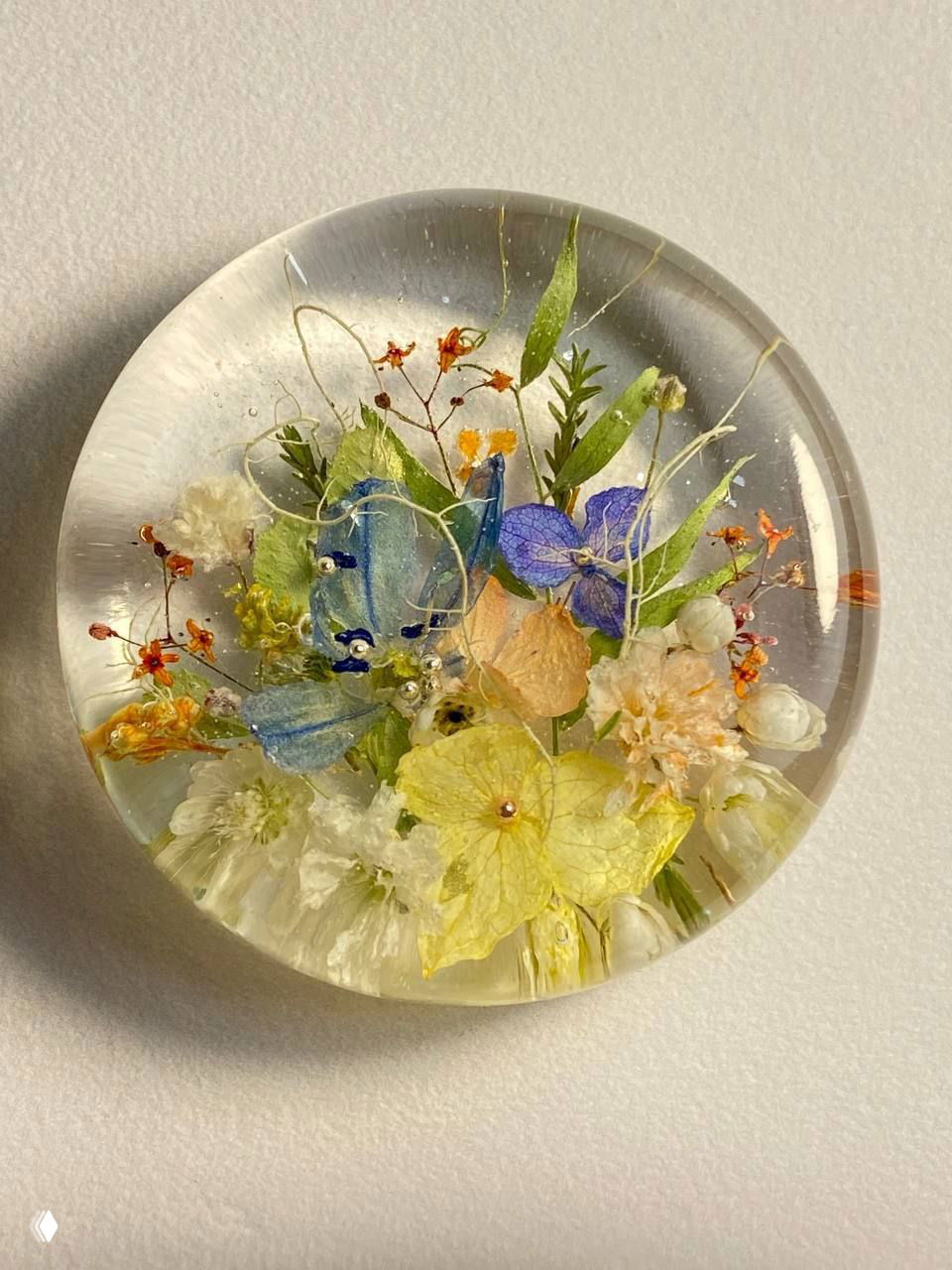 Крупный план эпоксидной медальонки с миниатюрными яркими цветами, бусинками и зелёными акцентами на светлом фоне.