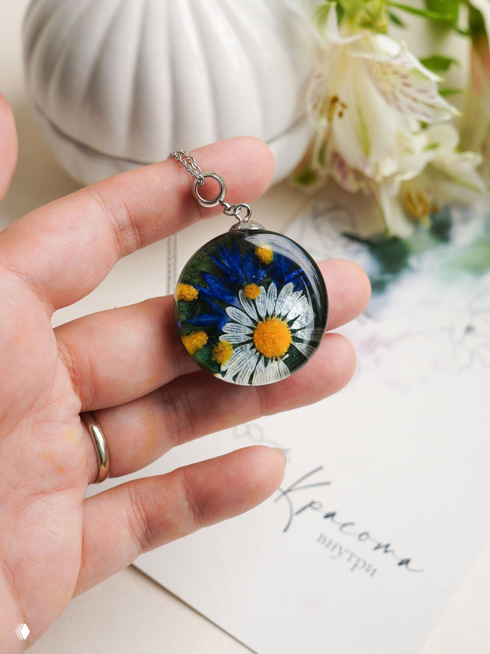 Крупный план круглой подвески из смолы с ромашкой, васильком и шариками мимозы; серебристая цепочка, изделие в руке.