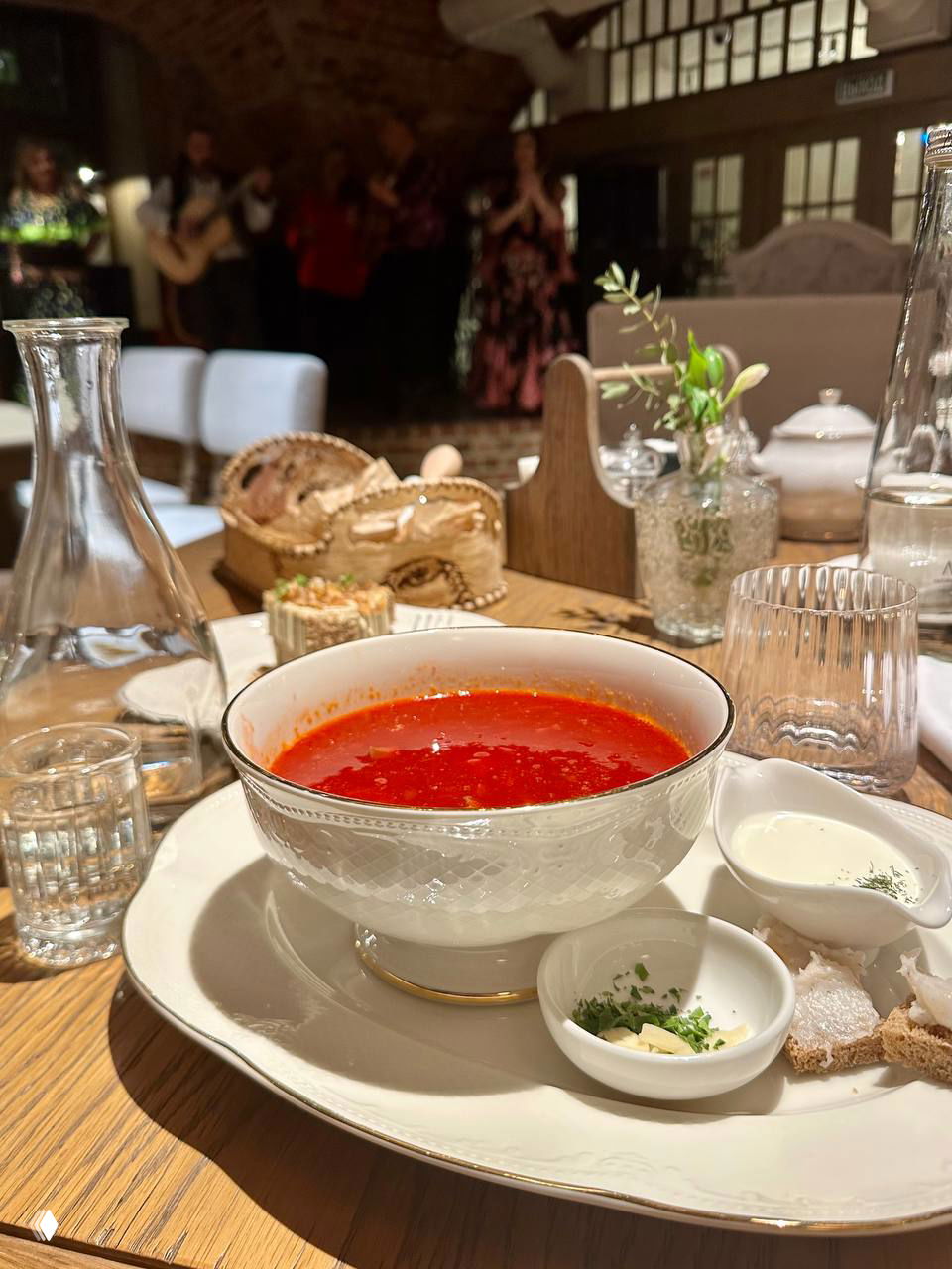 Тарелка с ярким борщом, небольшие блюда с добавками и рюмка на деревянном столе; на заднем плане размытая сцена с музыкантами.