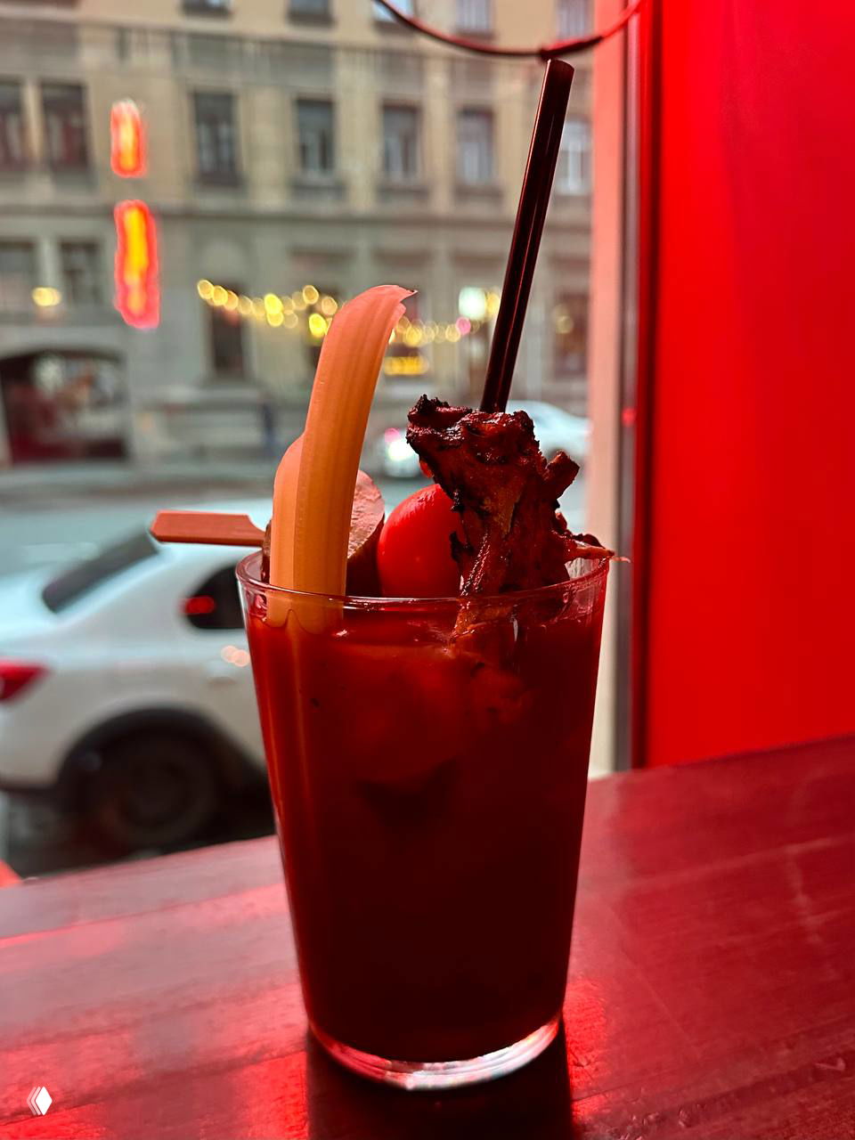 Стакан Blood Mary с бананом и мясной шпажкой на подоконнике; в окне видна улица Петербурга и красная атмосферная подсветка.