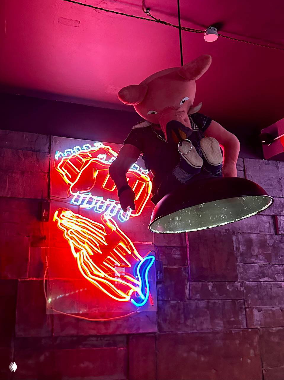 Неоновая вывеска и большая мягкая игрушка-свинья под потолочным светильником в интерьере бара с красной и синей подсветкой.