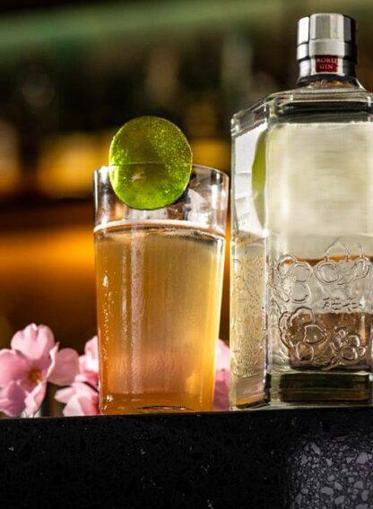 Стакан с янтарным коктейлем и ломтиком лайма рядом с декоративной бутылкой; акцент на подаче и эстетике бара в тёплых тонах.
