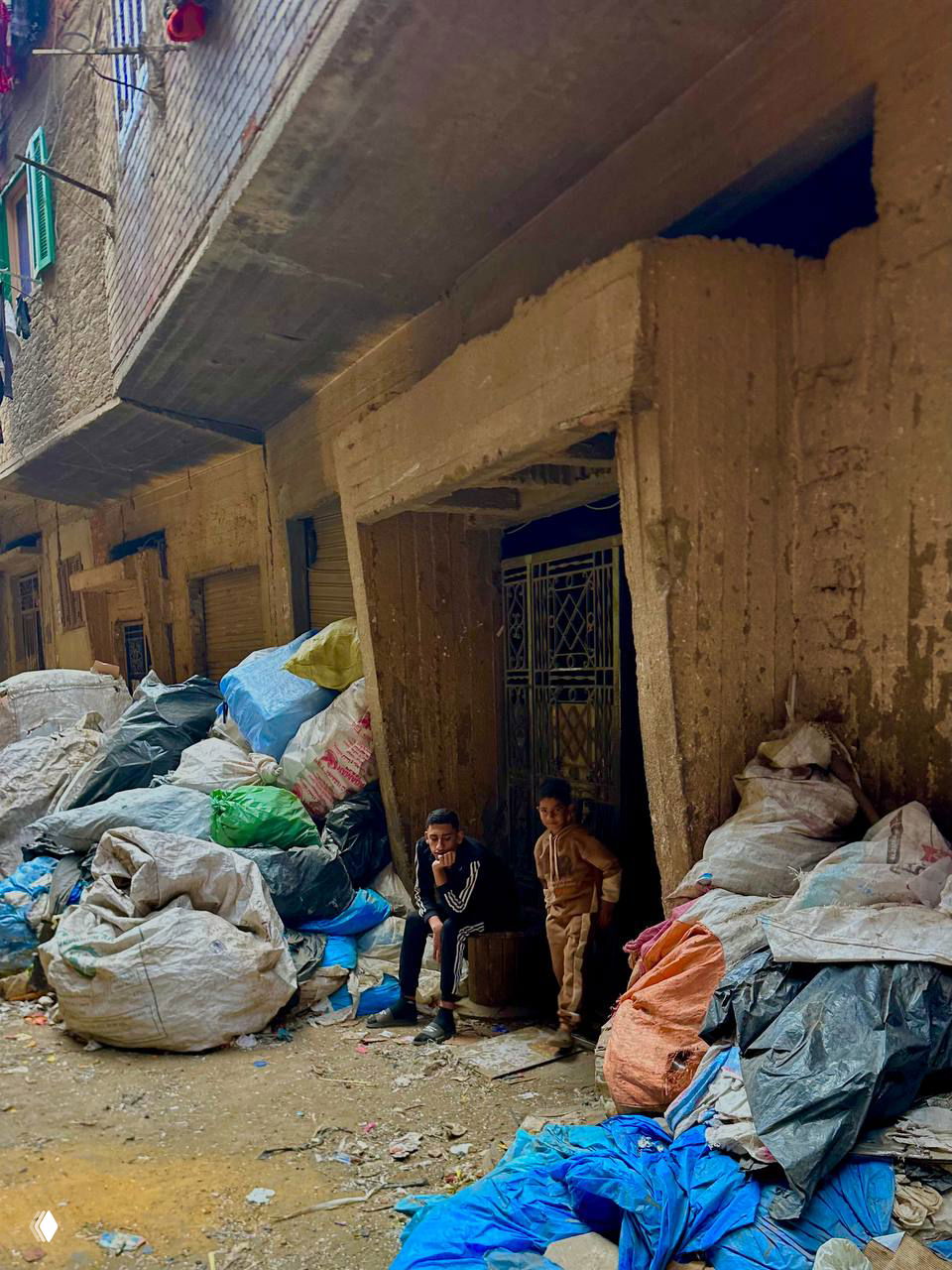 Узкий переулок, заставленный мешками с мусором; у дверей сидят двое мальчиков — сцена повседневной жизни в квартале переработчиков.