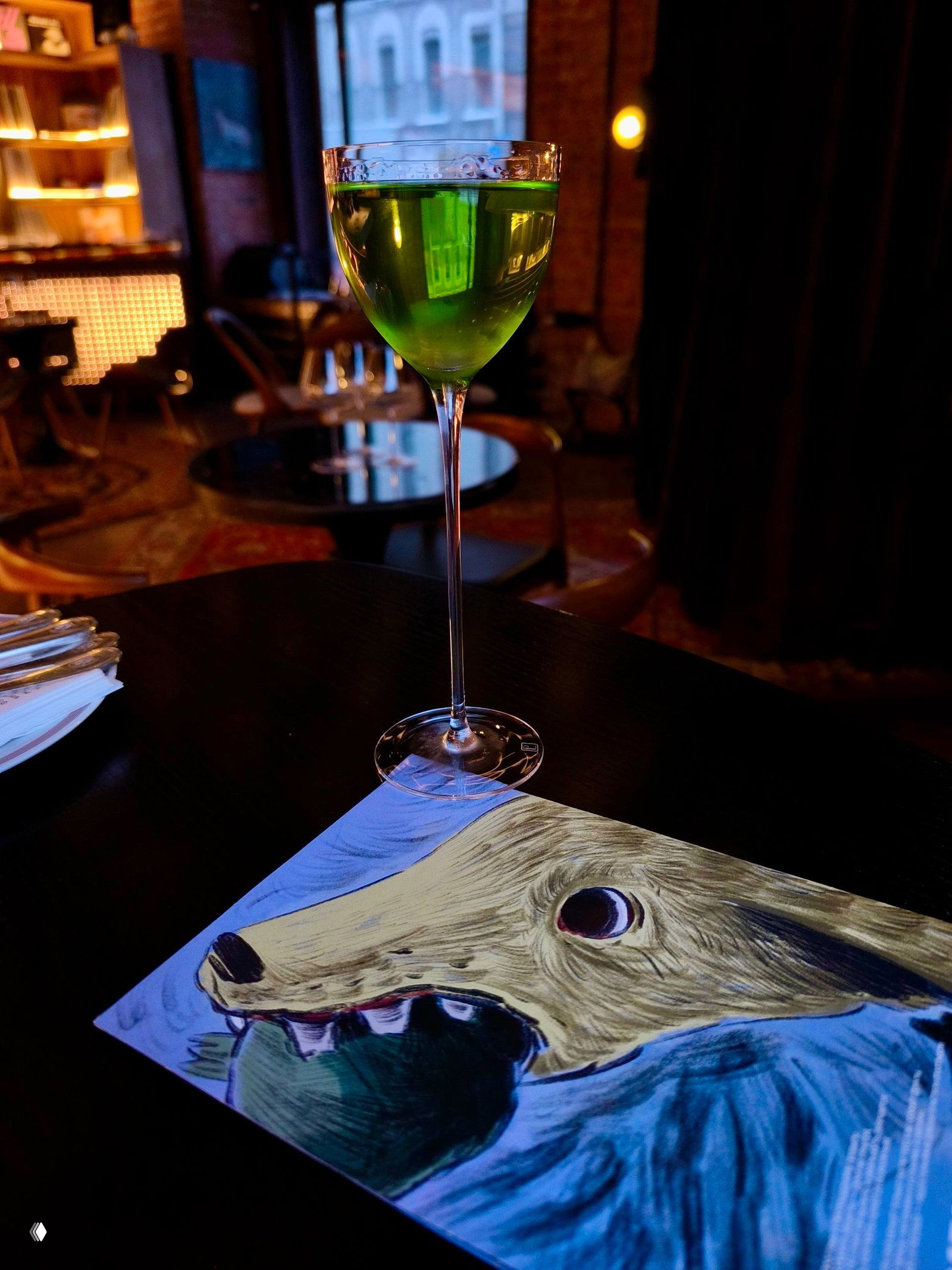 Высокий кадр зелёного коктейля в тонком бокале на фоне приглушённого интерьера и иллюстрации с изображением пьяной собаки