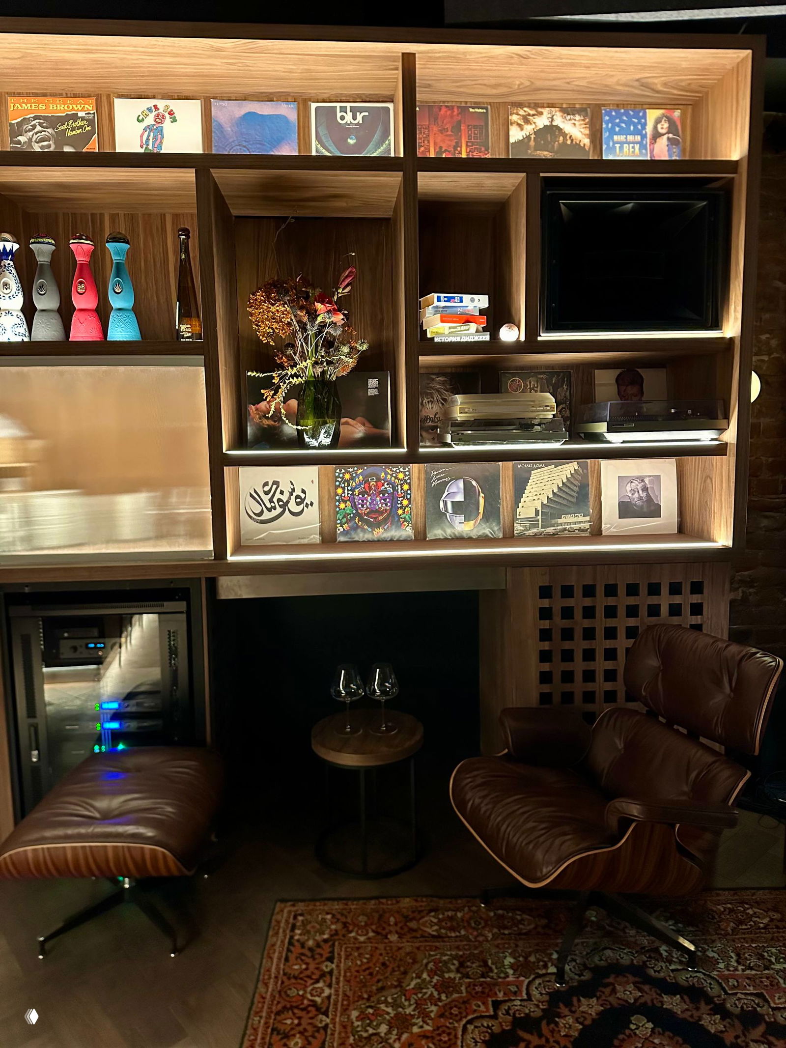 Уютный угол с креслом и оттоманкой рядом с барной стойкой; на полках винил и декоративные предметы, создающие лаунж‑атмосферу