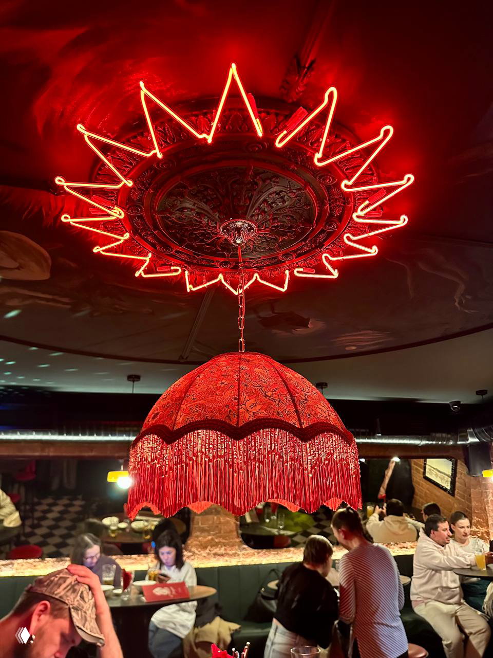 Низкий потолок и большая красная люстра с бахромой, неоновая декоративная подсветка в форме венца, общая сцена зала с гостями и барной атмосферой в тёплых оттенках.