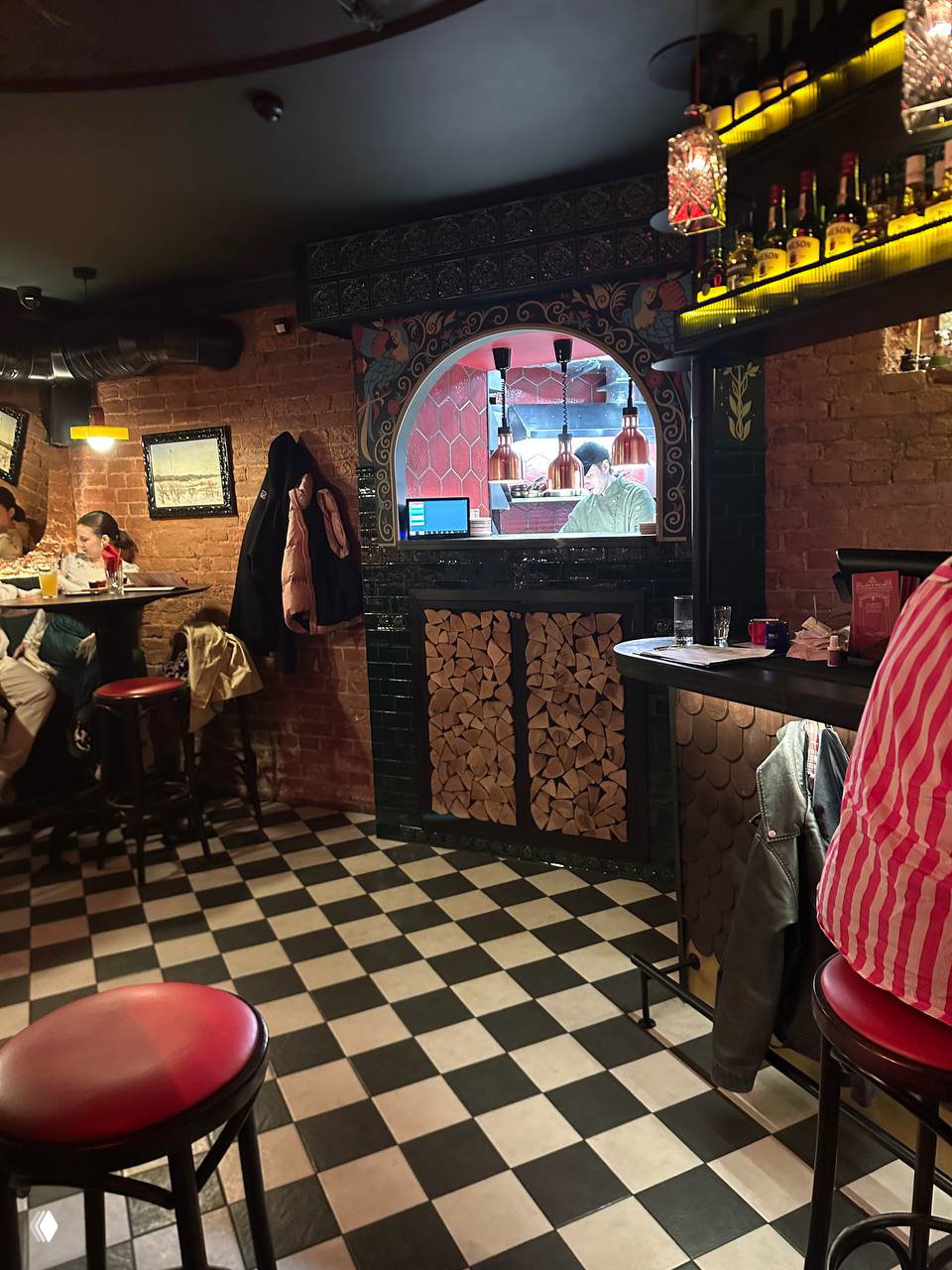 Барный зал с чёрно‑белой шахматной плиткой, барной стойкой, арочным окном на кухню и декоративными поленьями в оформлении — вид обращён к гостям, сидящим за столиками.